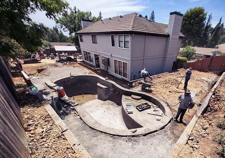 Gunite pool shell under construction in a Sacramento backyard, showing kidney-shaped excavation and raised spa before plaster