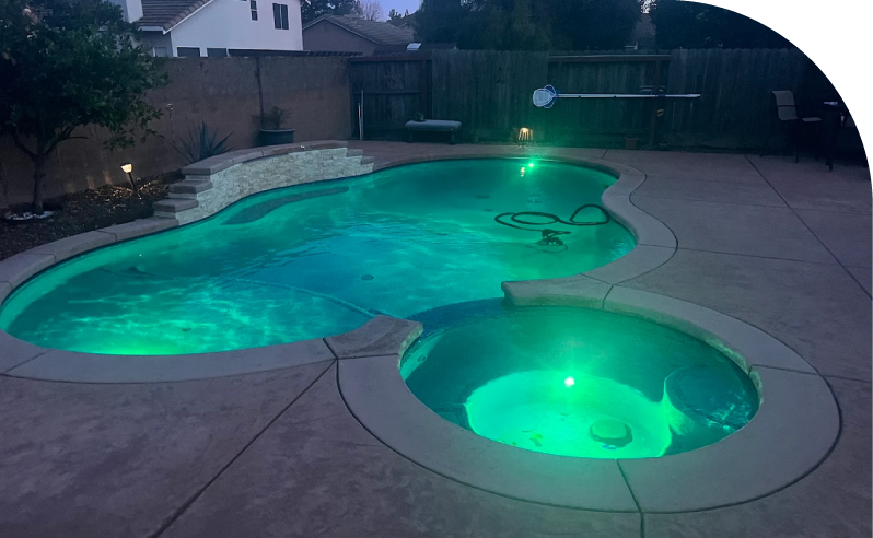 Custom kidney-shaped pool and raised spa with green LED lighting in a Sacramento backyard at dusk by trusted pool builders