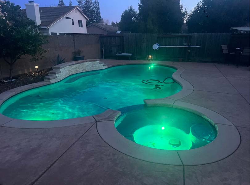 Kidney-shaped pool and raised spa with green LED lighting in a Sacramento backyard at dusk