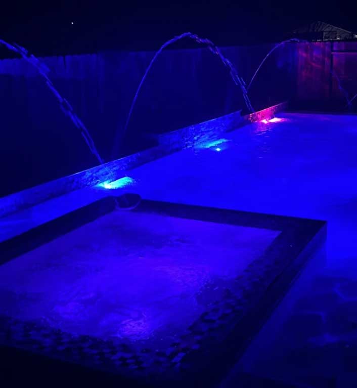 Sacramento pool with blue LED lighting and arching deck jets over a raised spa at night